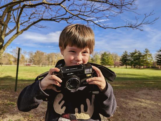 Male Human child looks happily at the viewer, a vintage Minolta camera in his hands.