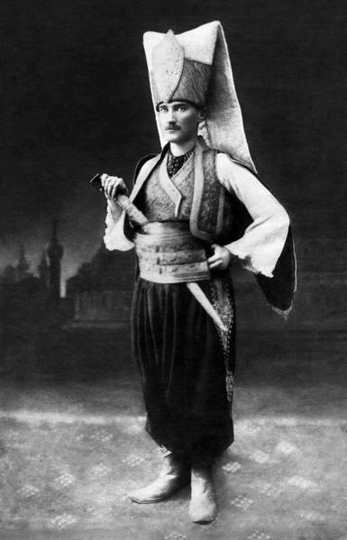 This is a photo that shows Atatürk in a Janissary uniform.

He wears a large headpiece of folded white cloth with a large red rim that stands a good 50cm high. It bears a golden signet on the forehead.

The outfit comprises a white flowy shirt under a hold and red vest with much embroidery.

A large cumberbund is wrapped around the waist in gold red and green.

The trousers are a harem style in a simple coarse black. A sword on placed inside the cumberbund.

Atatürk is in profile, in front of an oriental screen in a photo studio.