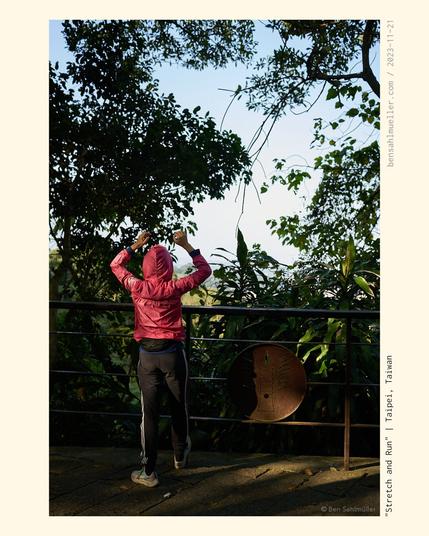 A woman in jacket, stretching with her arm lifted high while she looks down through a dense jungle onto Taipei.