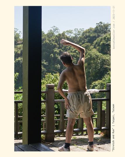 A man stretching while looking onto a forested hillside. He is wearing light shorts and no shirt.