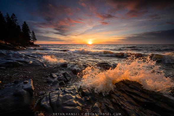 A photo of the sunrising over a large body of water. A wave illuminated by a wave splashes up on the black basalt shore. The clouds in the sky are red from the sunrise.