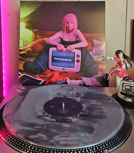 A clear vinyl record sits on a turntable. Behind the turntable, a vinyl album outer sleeve is displayed. The front cover shows a woman sitting on a bed with a tv screen between her legs. The screen says Digital Dreamscape. Around her are newspaper pages

To the right of the album cover is an anime figure of Yuki Morikawa singing in to a microphone and holding her arm out.