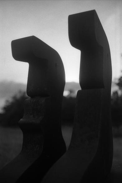 Ein Schwarzweißfoto des Kunstwerks "Er und Sie" von Franz Leinfelder, 2009: Zwei sehr großnasige Metallfiguren sitzen auf einem Metallsockel. Dahinter Sonnenuntergang.