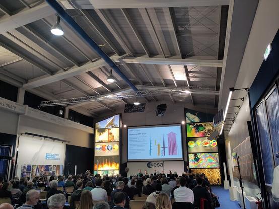 A conference hall full of people with a panel sitting along the top, a woman is presenting a slide. Colourful and very large satellite imagery of the globe decorates the sides.