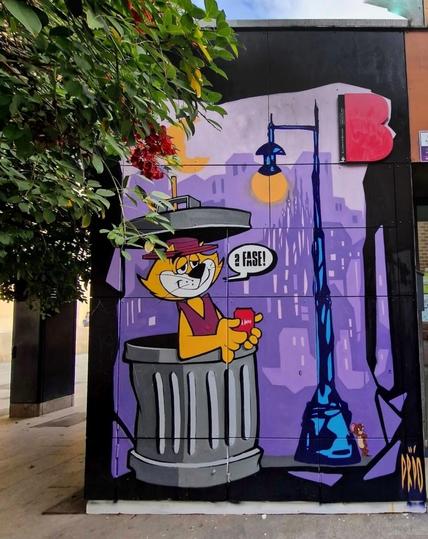 Streetartwall. A comic-style mural was sprayed/painted on the black exterior wall of a commercial building. A beige cat in a sleeveless red jacket looks out of a gray trash can with a smile. It has the lid on its head and is holding a red soda can. Next to her is a blue street lamp with a small gray mouse hiding at its base. The silhouette of a purple city at night can be seen in the background.
Info: The cat was created by the artist "a Fase !" (Edgard Vieira) from Spain and the mouse by PreoArt from Brazil.
