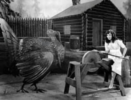 Black and white photo. A giant turkey looks on as a woman in a white dress sharpens her ax with a stone wheel