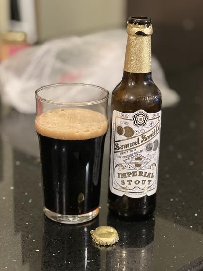 Samuel Smiths Imperial Stout in a pint glass. The brown bottle has a white label with black and gold lettering and a gold foil wrap around the upper neck and cap