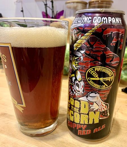 A glass of red ale and its can are on a pine wooden table. The can has an aggressive bloody unicorn, which probably reflects the color of this ale - dark amber red.