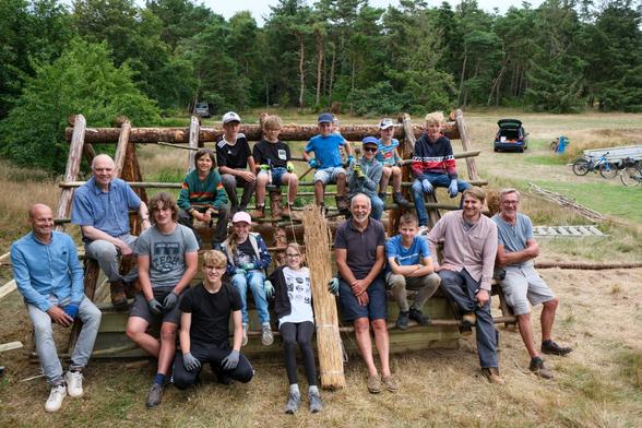 Eine Gruppe von Kindern, Jugendlichen und Erwachsenen sitzt auf den Dachbalken eines kleinen Holzhauses. Alle blicken hoch in die Kamera. Das Haus steht auf einer Waldlichtung.