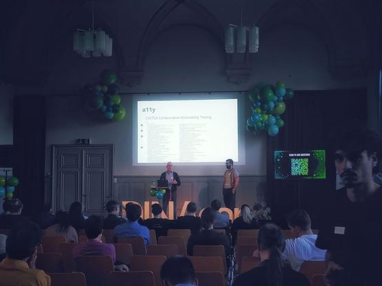 People sitting in a darkened conference room, watching a presentation on Accessibility testing. On the screen behind the two male speakers is a slide with the title "a11y". There are balloons is a QR code for audience feedback next to them. A man, a photographer himself, looks into the camera questioningly.