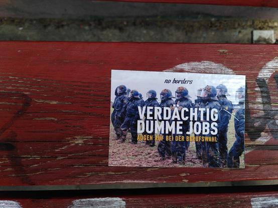 Auf einer rot lackierten Holzbank klebt an der Rückenlehne ein Aufkleber. Darauf zu sehen ist ein Foto von Polizist*innen in Riot Montur mit Helmen und Schutzausrüstung. Vermutlich ein Schnappschuss von der Großdemonstration gegen die Zerstörung Lützeraths am 14.01.2023. Die Cops stehen im Schlamm und sind an vielen Stellen verdreckt, als hätten sie ein paar Schlammbälle mit den Köpfen gefangen.

Das Bild wird wie folgt betitelt:
Verdächtig dumme Jobs. Augen auf bei der Berufswahl.
Oben steht noch etwas kleiner "no borders"