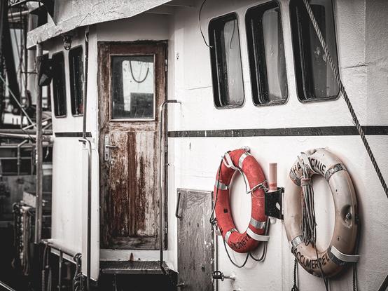 Ausschnitt eines Schiffes. Zu sehen ist eine abgenutzte Holztür, 2 Rettungsringe und drei aneinander gereihte Fenster.
