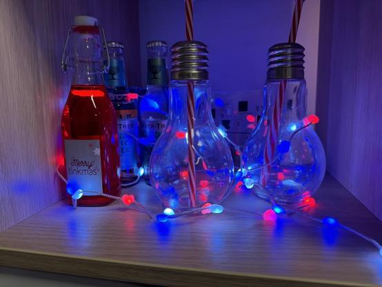 Die Twinkly Candies-Lichterkette mit Herz-Lämpchen liegt eingeschaltet in einem Regal mit Getränkeflaschen und Gläsern.