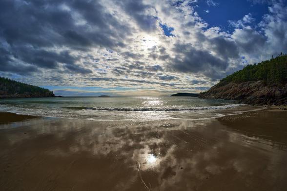 The sun is screened by banks of fluffy cloud in a deep blue sky. The scene is reflected in the wet sand. The sea is choppy and green and the the beach is banked by rocks and evergreen trees.