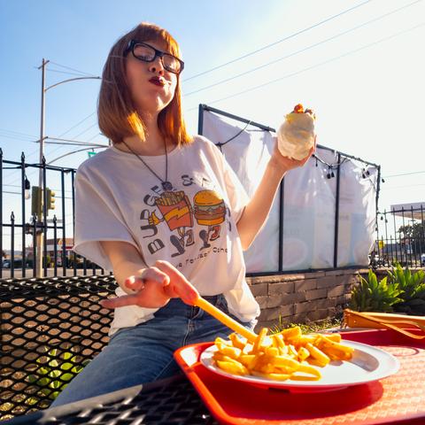 Photograph of Lety seated in the outdoor dining section of a restaurant. She is wearing a graphic tee and denim blue jeans. Lety's t-shirt features an anthropomorphic hamburger and carton of fries holding shoulders as they stroll and the text "Burgers & Fries, Made for Each Other." She calls this her Nona shirt. Lety's mouth is stuffed full with a bite of the burger in her left hand as she reaches for a french fry from the plate in front of her with her right.