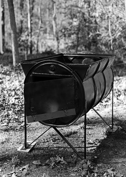 Black and white image showing the stove base of a small evaporator used for making maple syrup. It's made from a 55 gallon barrel that's been flared at the top to hold a pan. There's a door for putting in firewood, and it stands on simple metal legs.

It was made in Vermont by a local company (now defunct) called Ascutney Forge. I used it for a number of seasons, and it was rusty after years in storage. It's been freshly painted black, with an added coat of clear acrylic.