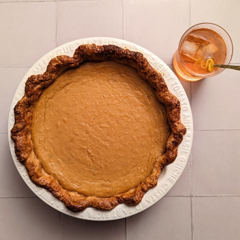 A close-up of a pie in its dish, sat next to a cocktail in a rocks glass, on a cream tiled background. The pie has a golden crust and pumpkin-orange filling. The cocktail is a orange-brown liquid, on ice, with a lemon peel garnish.