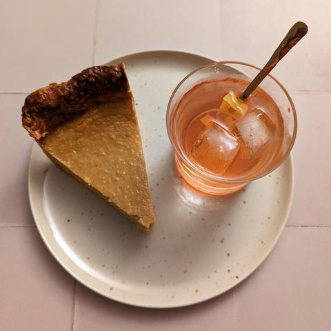A slice of the same pie sitting on a speckled cream plate next to the same cocktail, on the same cream tiled background.