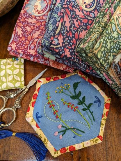 Brauner Holztisch. Darauf ein großes Stoff-Hexagon aus blauem Leinen mit Blütenstickerei in der Mitte. Gerahmt ist es mit gelbem Stoff, der grüne Ranken und rote und weiße Blüten trägt. Links daneben eine kleine silberne Nähschere  mit blauer Tassel. Daneben ein kleines, grün-weißes Kissen mit Näh- und Stecknadeln. Im oberen Bilddrittel sind gefaltete Stoffstücke mit floralen Mustern zu sehen.