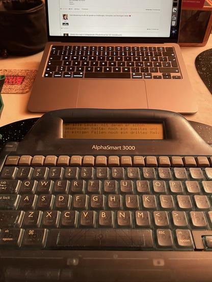 Ein AlphaSmart 3000 im Vordergrund, dahinter ein MacBook Air. Der AlphaSmart ist mehr oder weniger eine Tastatur mit einem 4-zeiligen LCD in einem halbdurchsichtigen grünen Plastikgehäuse. Dieses Modell wurde erstmals im Jahr 2000 vorgestellt, lange bevor der Begriff „distraction free writing“ erfunden wurde.