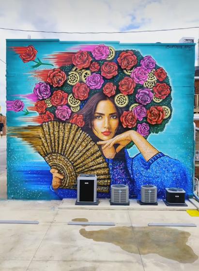 Streetartwall. Painted and animated building facade. The mechanical system uses 12 motors to operate the painting. The colorful mural of a young woman with a Hand fan is sprayed/painted on a modern three-story building. The background is blue. On it is a young dark-haired woman wearing a voluminous arrangement of red and purple roses with gold gears in between as a headdress that takes up half the wall. She is wearing a blue woolen sweater and holds one hand pertly under her chin. In the other hand she holds a large, beige-patterned Hand fan. The picture is painted on the left side as if it had been blown away by the wind.