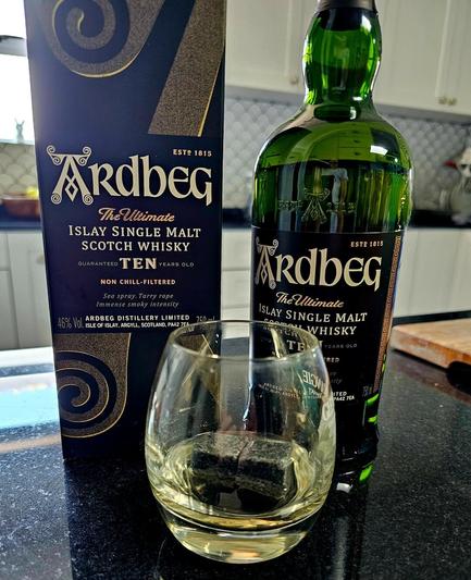 A bottle of Ardbeg 10 Year single malt whisky, standing next to it's box, with a glass in the foreground that has a little whisky in with two whisky stones.