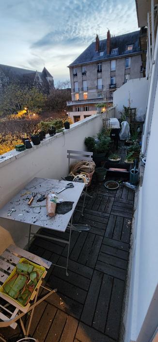 Au crépuscule, un balcon filant où régnent le bazar et les poignées de terre éparses. Au premier plan, une coupelle avec des tessons de poterie posée sur une chaise ; une petite table couverte d'outils, de godets, de terre et de saletés ; derrière, un amoncellement de bacs, pots et plantes au sol.