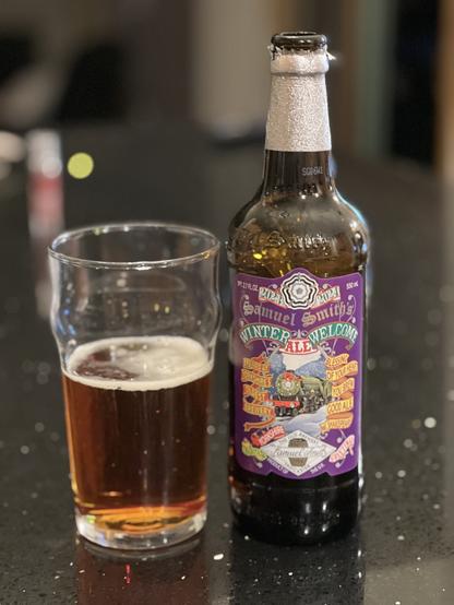 Samuel Smith’s Winter Welcome Ale. A tall brown bottle adorned with a silver foil collar. The label is purple with red yellow and green banners declaring “Brewed at Yorkshires oldest brewery” and “Blessing of your heart, you brew good ale. W. Shakespeare”