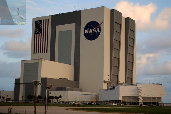 An outdoor, morning photograph of a tall white and gray rectangular building, with a tall extension from one face, and a bent, white section building next to it.
The large building has a large US flag painted on vertically on one side and the blue NASA meatball logo on the other.