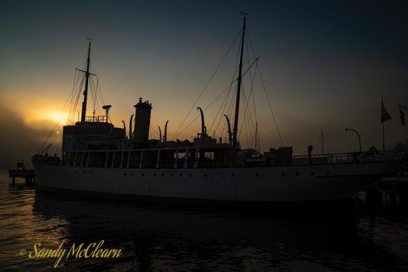 Ship in silhouette from a rising sun partially obscured by the background fog.