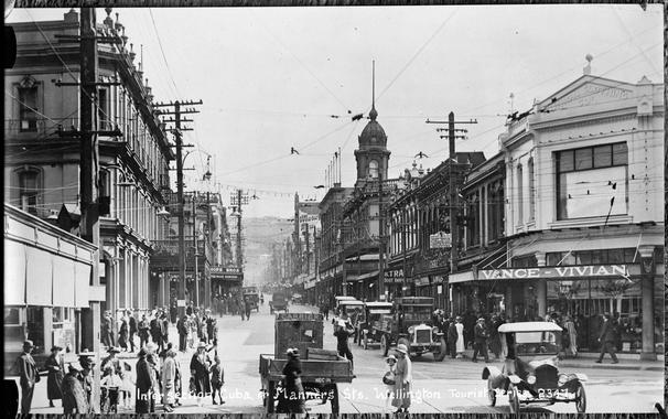 Black-and-white photo: Intersection of Cuba Street and Manners Street, Wellington, 1920s? Description: Looking up Cuba Street, Wellington from the Manners Street intersection, showing the premises of Vance Vivian on the corner (right); boot importers; tram stop sign on power pole; Te Aro House and other buildings (right to rear); on left various buildings then Royal Oak Hotel, Hope Bros the Value Mercers; crowds of pedestrians both sides of street and crossing road; cars and truck (foreground to rear); policeman controlling traffic (centre). Citation: Auckland Libraries Heritage Collections 35-R1763. https://kura.aucklandlibraries.govt.nz/digital/collection/photos/id/275017