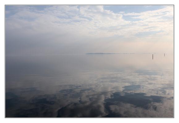A calm blue/white sky mirrored in the sea. In the distance an island, nearer two seamarks.
