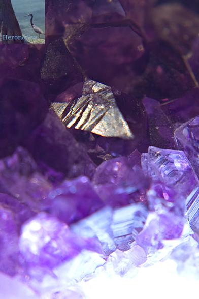 A vertically-oriented photograph of a purple amethyst crystal. At the top of the frame and furthest away is a single, flat peak of crystal reflecting the light. Below and in front of it is jumbled crystalline chaos.