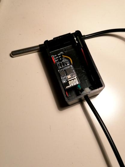 A ESP-01 based temperature sensor. The DS18B20 is external stored in silver case left above. Inside parts of the breakout board are visible including some wires. On top of it a ESP-01 is placed. The board has label for the the pins.
On the upper right there is a micro-USB port glued to the side. Almost not visible the step-down module which is glued to side below as well.