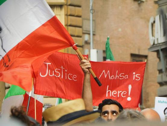 pictures taken at the march i mentioned in the caption.   two red banners here:  "Justice"  and  "Mahsa is here!"  written in white on them .   a man is holding the iranian flag  in front of them