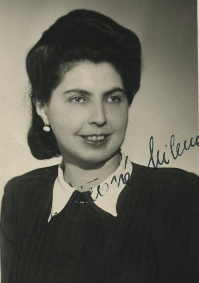 Portrait photography of a woman. She has very high up dark hair. An earring can be seen in the visible ear on the left. Her smile reveals her front upper teeth. The woman is wearing a dark blouse with a white collar. A handwritten signature can be seen on the photograph.