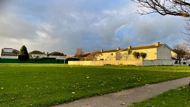 Across a suburban common green, the back of terraced houses are spot lit by intense golden light against a moody and cloudy sky.