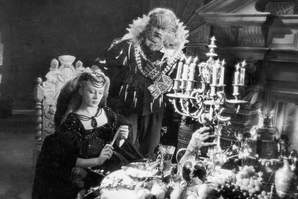 The youngest daughter seated at a lavishly set table with the Beast standing behind her. As magical as the story are the production and set design by Christian Bérard, Lucien Carré, and René Moulaert. They breathed a soul into almost everything, including the candelabras.
