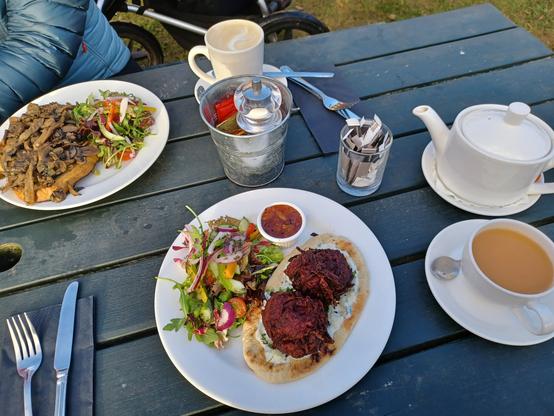 A picnic bench laid with tea and coffee, a plate of mushrooms on toast with salad garnish, and a plate of beetroot bhaji with flatbread, salad and tzatziki.