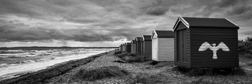 A grainy black and white image of the famous Findhorn beach huts. These are wooden huts painted in different colors set right at the high water mark. The different colors show as different shades of grey, with a large white gull painted on the side of the nearest hut. The beach is made up of large stone shingle, waves crash onto the shore on a fierce windy day. Grey skies complete the scene.
