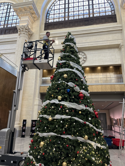 A man stands high up on a hydraulic lift, putting decorations on the top of a truly enormous artificial Christmas tree, in the sunny Beaux-Arts lobby of the Senate of Canada Building, aka the grand old Ottawa train station.