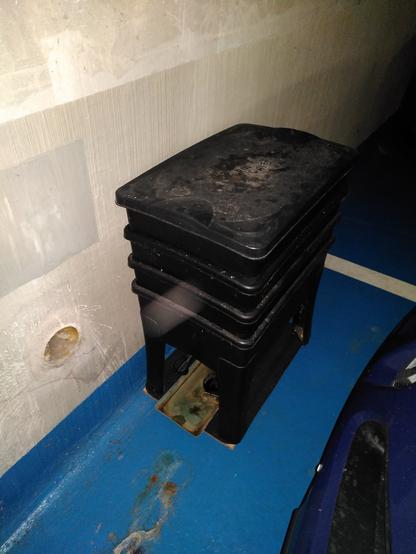 Bac lombricomposteur adossé a un mur de parpaings brut sur un sol peint en bleu on devine le capot d'une voiture juste à coté