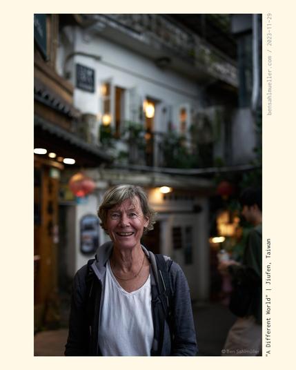 A portrait of a woman, wearing a white shirt and a grey jacket and looking joyfully into the camera. The image is taken spontaneously in the streets of Jiufen. Some of the houses can be seen warm and blurred in the background.