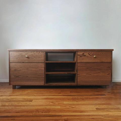 A red oak console. The center has open shelves while the left and right have 1 drawer and 1 door below that. It sits against a white wall on red oak floors.