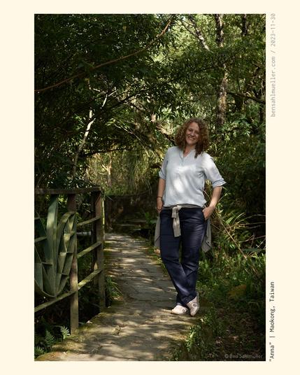 A curled young woman standing on a stone path on Maokong hill close to Taipei. The sun is shining through the leaves around her. She is smiling warmly.