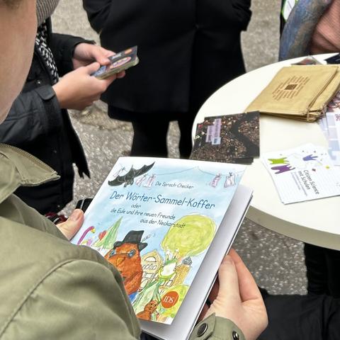 Eine Person hält das Buch der Sprach-Checker "Der Wörter-Sammel-Koffer" in Händen. Um einen runden Stehtisch herum (mit Informationsmaterial zu Citizen-Science-Projekten) sieht man weitere Personen. Die Personen und der Tisch sind nur im Anschnitt zu sehen.