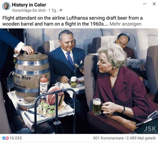 Ein servierwagen im Flugzeug mit Bierfass und Schinkenhäger Flasche, rechts davon sitzen 2 Männer und essen mit einem biet dabei, in der Reihe davor dreht sich rin Frau mit Bier um und schaut den Wagen an.