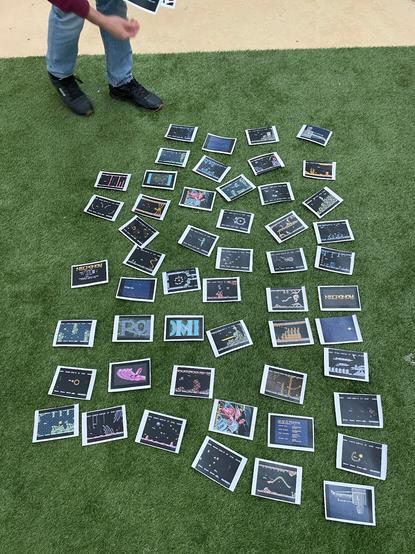 Printed screenshots of a video game lay orderly on a mat of fake grass. A person is moving towards them in order to interact with them.