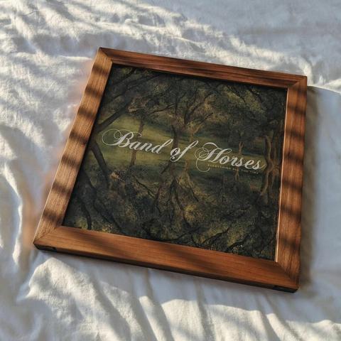 A walnut frame with a Band of Horses record in it laying on a white sheet with the sun casting shadows across it.