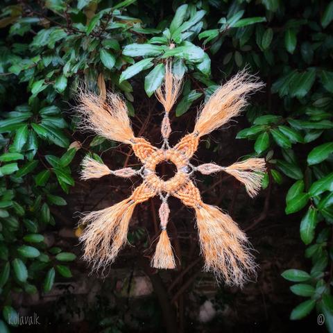 Photo couleur au format carré 

On voit une étoile à 8 branches faite de corde de jute détressée et fils noirs-dorés autour d'un cercle métallique recouvert (le tout de récupe) 
Elle est accrochée dans les feuilles vertes d'un oléastre.